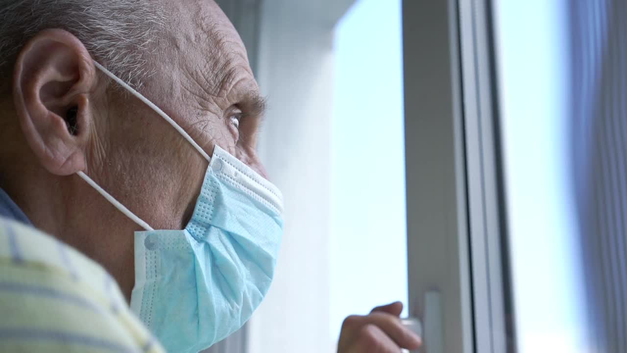 一位戴着蓝色无菌口罩的老人站在白色塑料窗前视频素材