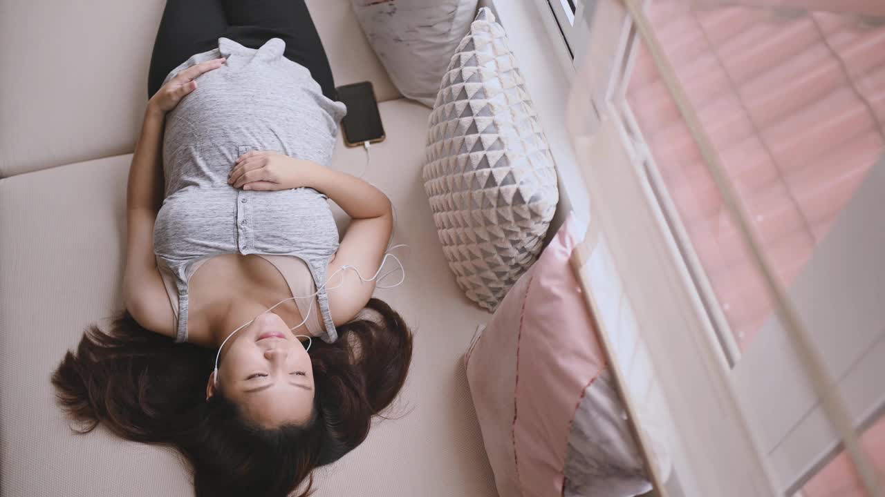 正上方是一名亚洲华裔孕妇，她躺在白天的床上，享受着智能手机播放的音乐视频下载