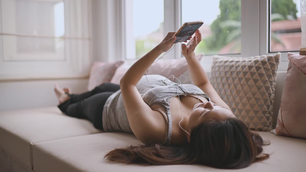 一名亚洲华裔孕妇躺在白天的床上，放松地享受着智能手机播放的音乐视频下载