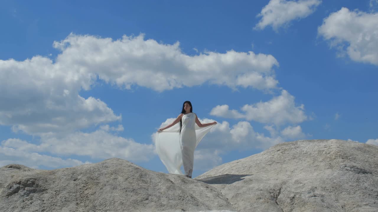 山顶上站着一位美丽的黑发长发女子，她穿着飘逸的白色衣服，映衬着天空。天使存在的幻影。4k 50fps慢动作视频下载
