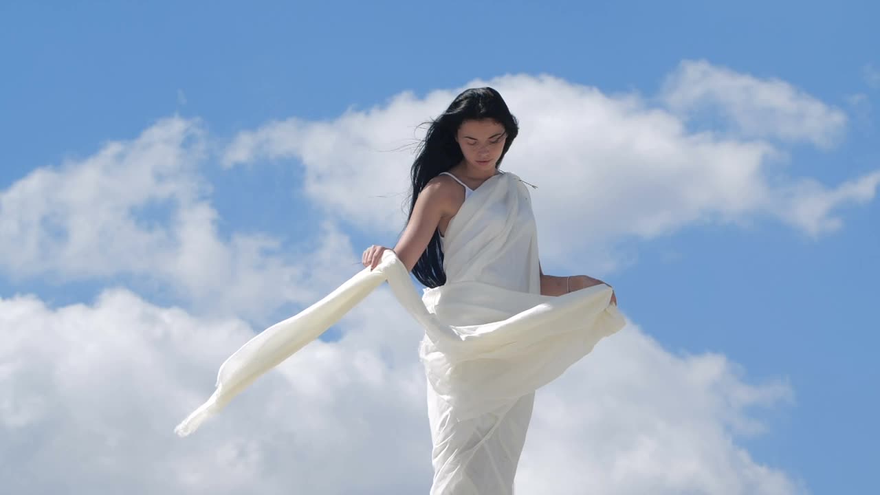 山顶上站着一位美丽的黑发长发女子，她穿着飘逸的白色衣服，映衬着天空。天使存在的幻影。200帧/秒的慢动作视频下载