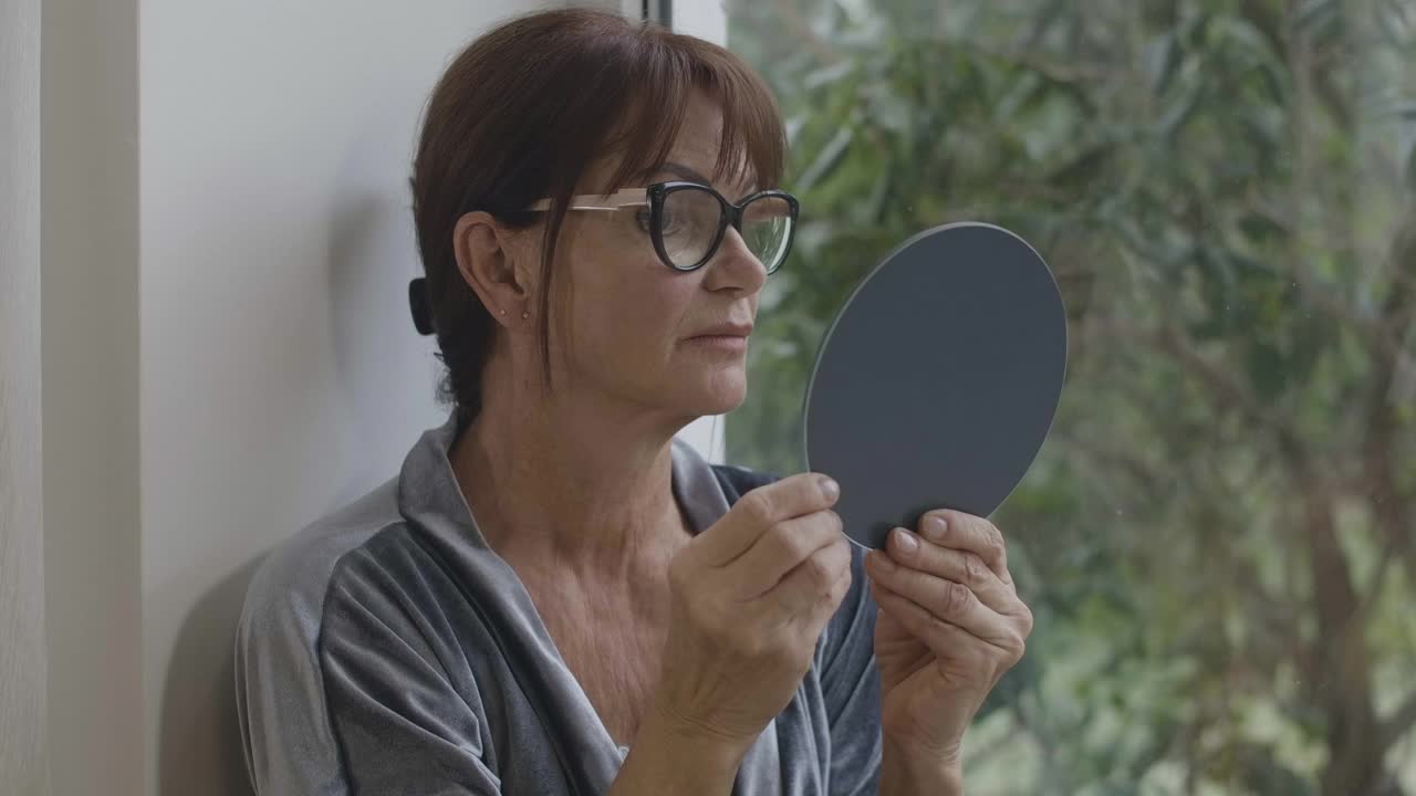 近距离的肖像自信的资深妇女在眼镜看反射在手镜。时尚的白人退休人员检查皱纹。老龄化和幸福的概念。电影4k ProRes总部。视频素材