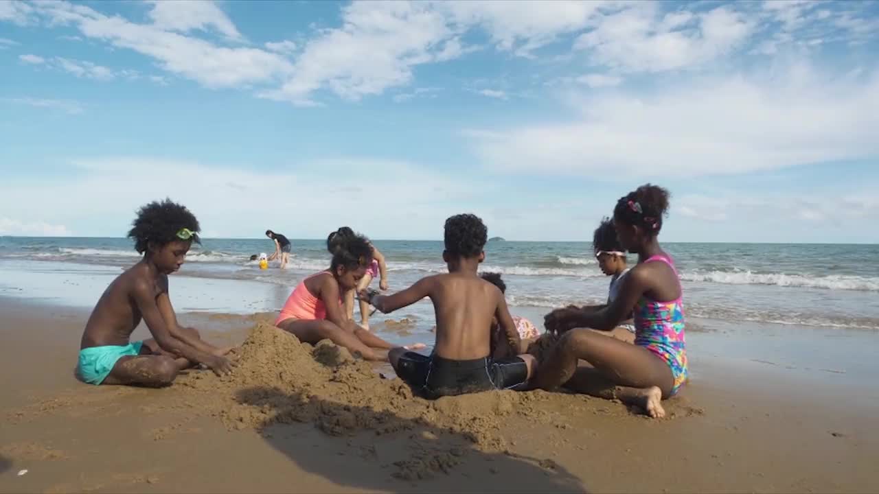 一群非裔美国儿童在有云、蓝天和大海背景的海滩上挖沙子视频下载