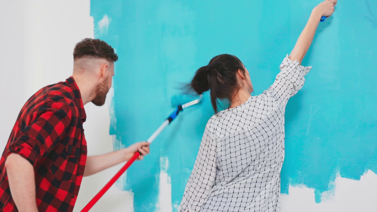 美丽的夫妇装饰他们的新公寓和胡闹。夫妇俩正在用蘸有浅蓝色颜料的滚筒刷墙。他们很快乐，玩得很开心。家里装修视频下载