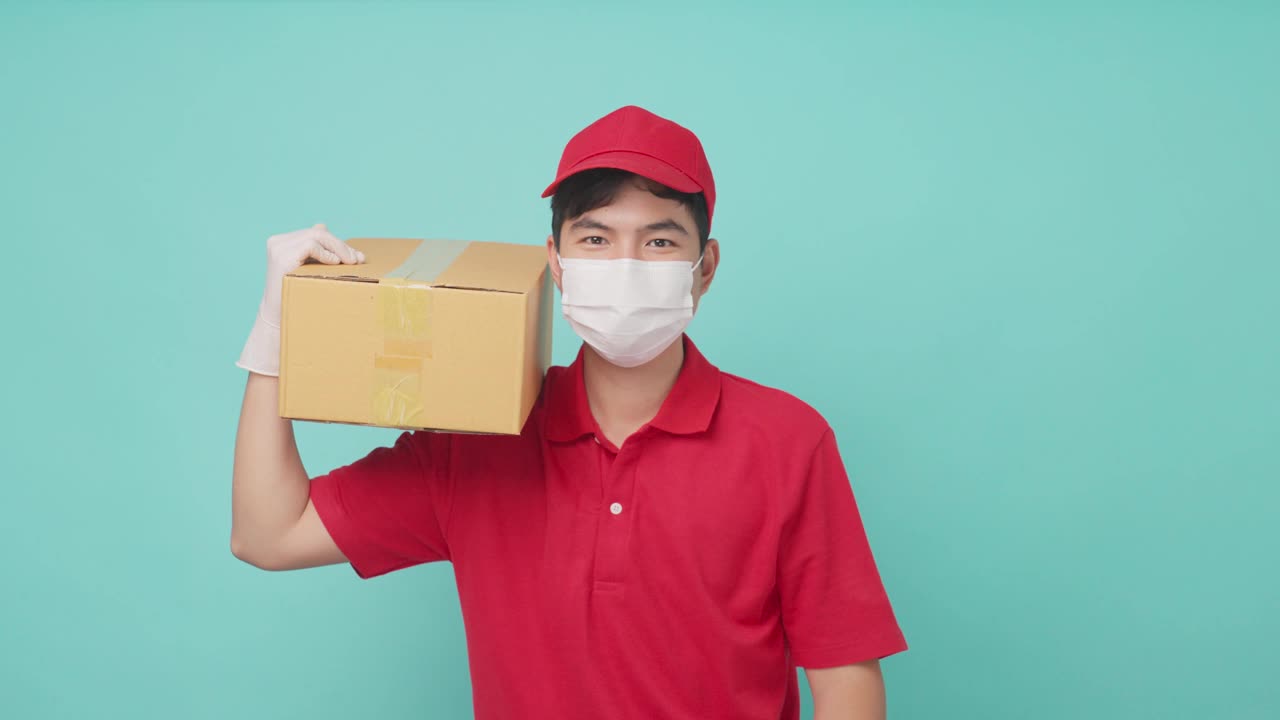 孤立的背景下，身穿红色制服的亚洲快递员戴着面具，肩上扛着一个中等大小的盒子包裹，准备递送给客户。男邮差竖起大拇指，面带微笑。视频素材