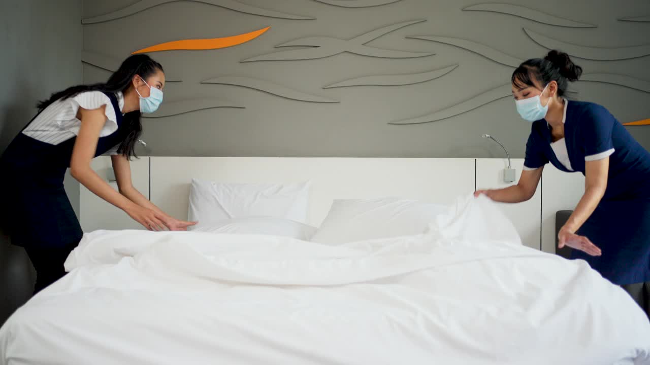 两个年轻的女仆带着医用口罩在酒店房间的床上更换床单。视频素材