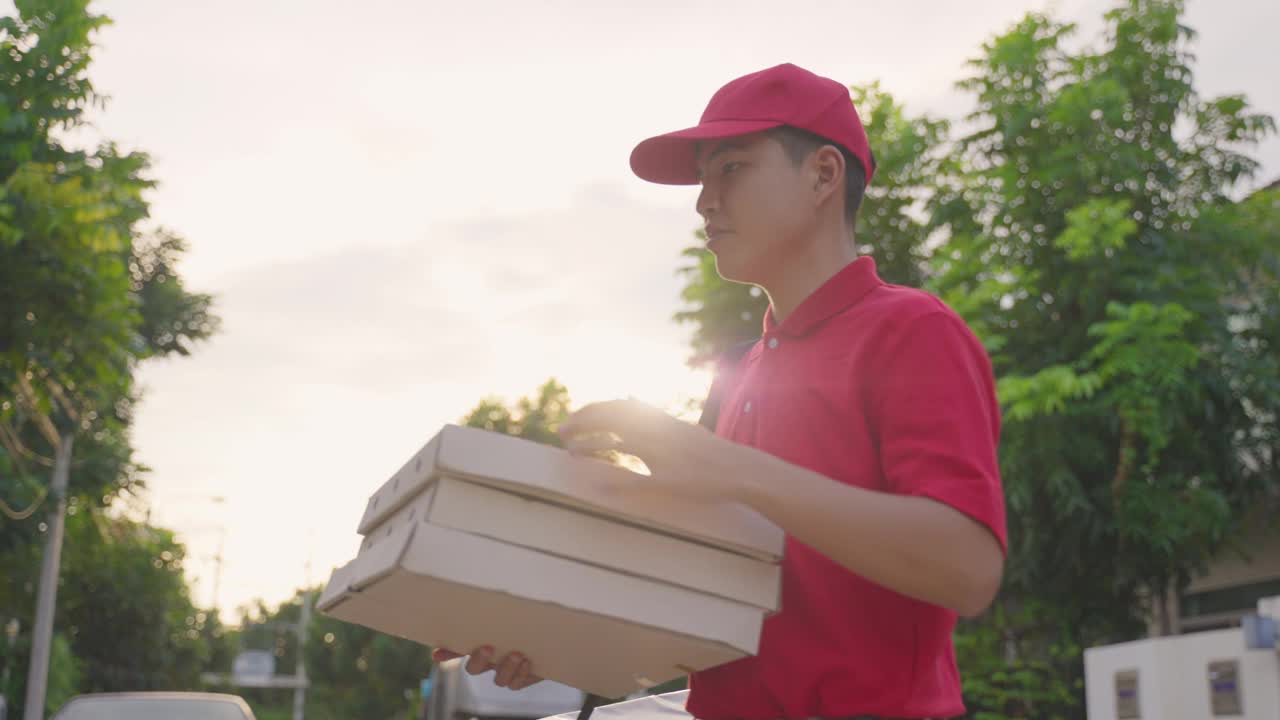 亚洲送货男工人穿着红色制服搬运食物，披萨给年轻漂亮的女顾客在房子前面。邮差和快递杂货配送服务理念视频素材