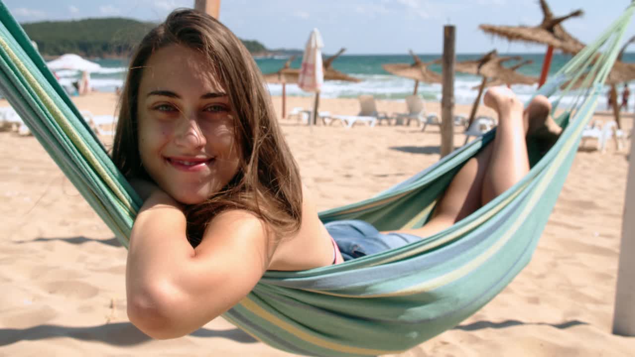 一位年轻女子躺在吊床上，享受着海滩上夏日的阳光。暑假假期旅行概念。4 k慢动作视频下载