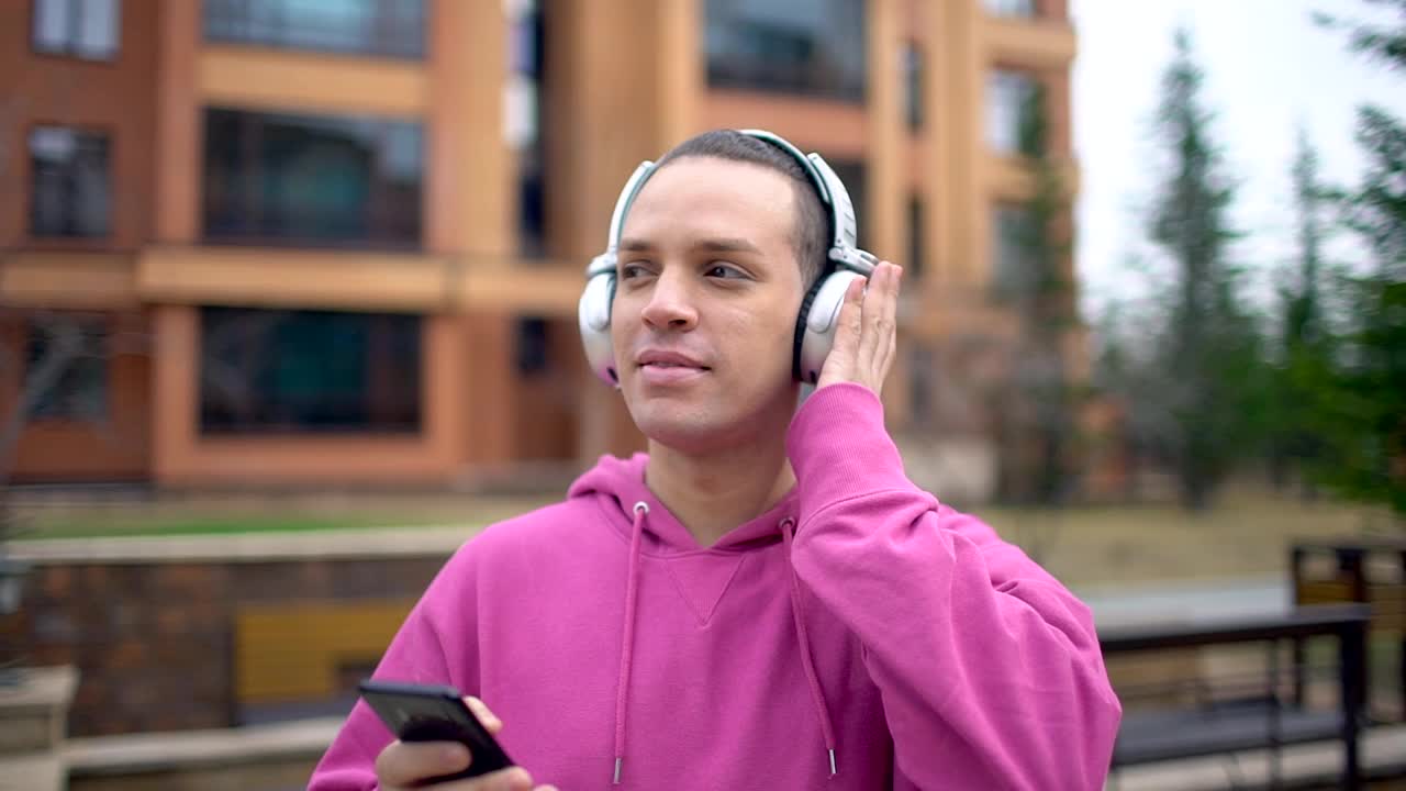 一个年轻人在街上戴着耳机。手持智能手机的男子走在城市街道上。年轻的白人喜欢音乐。英俊的男人用智能手机和耳机听音乐视频素材