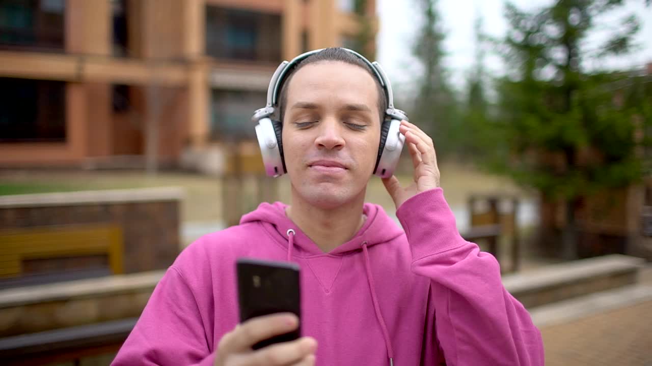 人，音乐，科技，休闲和生活方式-快乐的年轻潮人戴着耳机和智能手机走在城市的街道上。年轻有吸引力的男子在城市背景下听音乐耳机的肖像视频素材