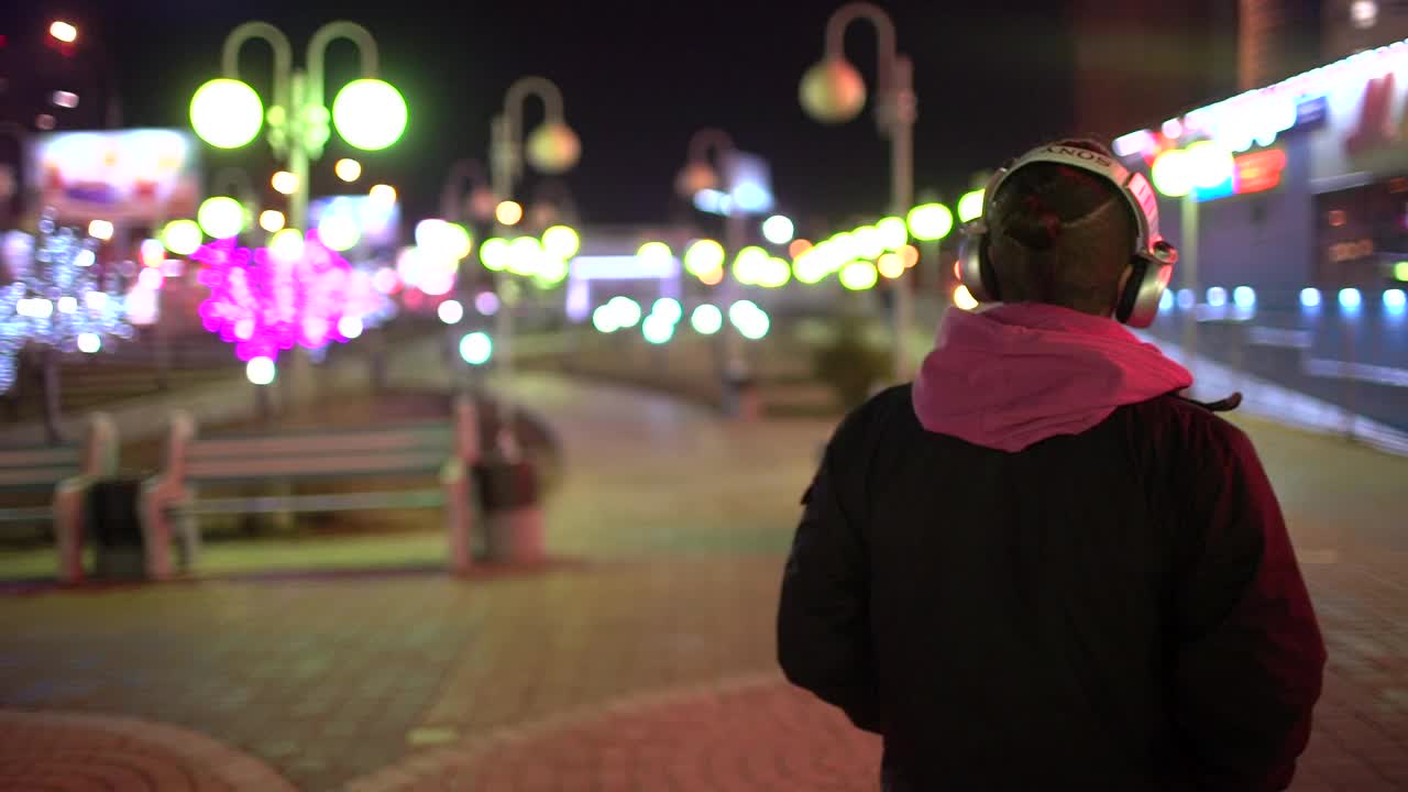年轻男子的肖像戴着耳机，而走在街道上在晚上的城市和开始听音乐。戴着耳机走路的潮人视频素材