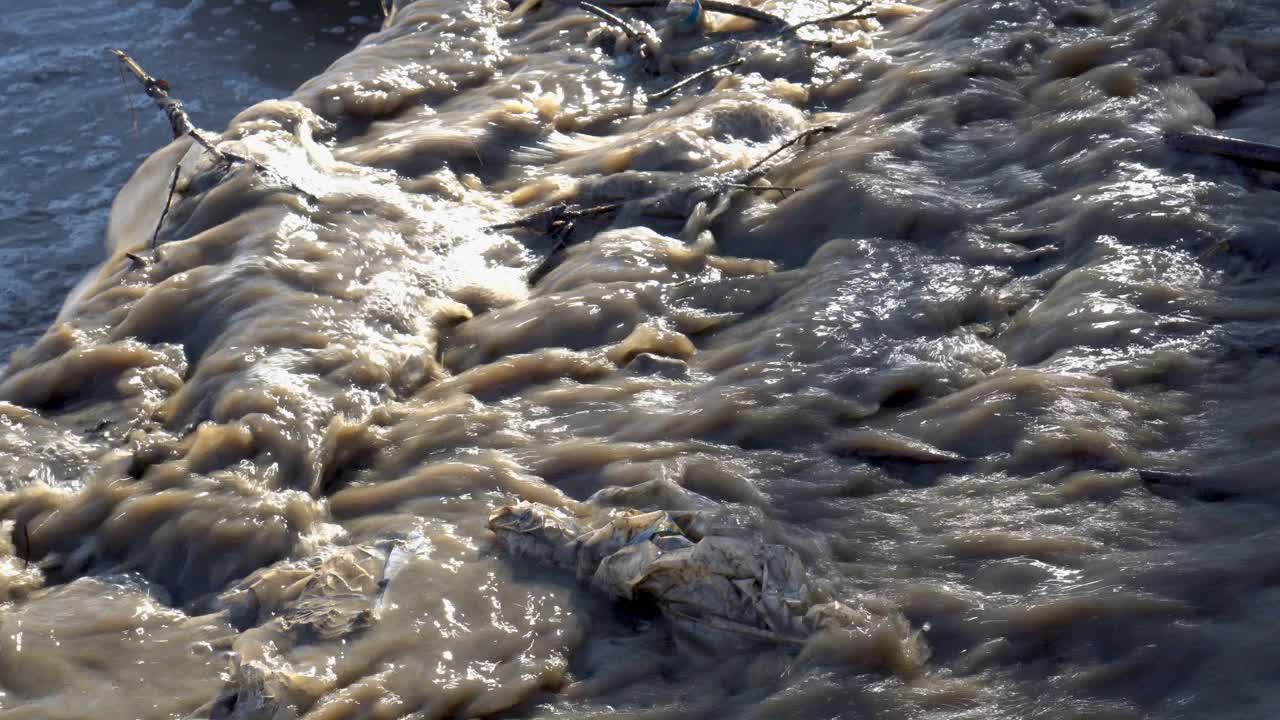 一条肮脏的、褐色的水带着碎片、树枝和塑料流进河里。环境污染的概念。特写镜头。4 k。视频素材