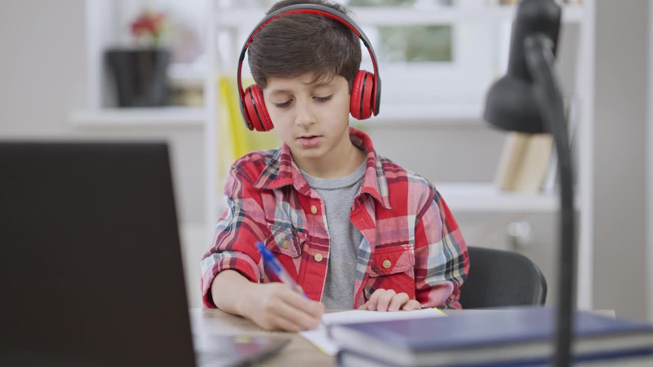 一幅兴高采烈的中东男孩的肖像，他戴着耳机坐在室内桌子旁唱歌，用慢动作做手势。粗心的小学生一边做作业一边戴着耳机听音乐。视频下载