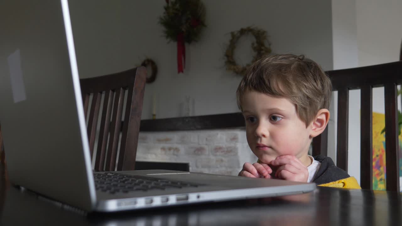 蹒跚学步的孩子坐在家里的桌子上看笔记本电脑视频素材