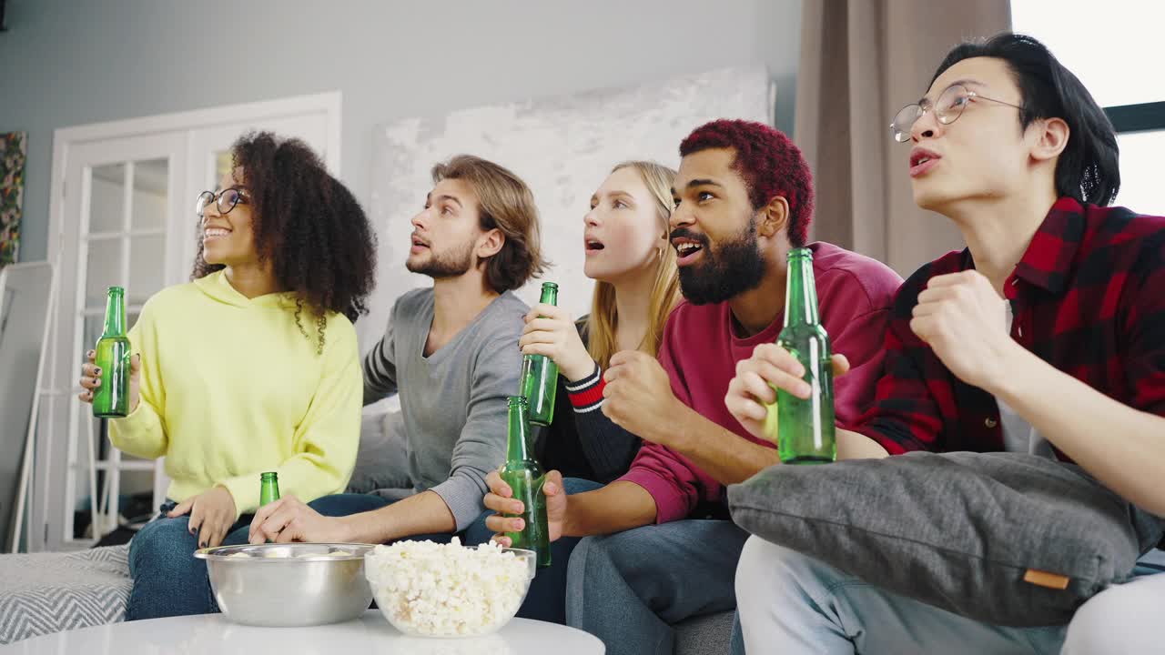 快乐的混合种族的朋友有乐趣的室内看电视体育一起，喝啤酒，为团队欢呼。庆祝自己喜欢的球队胜利的一群人视频下载