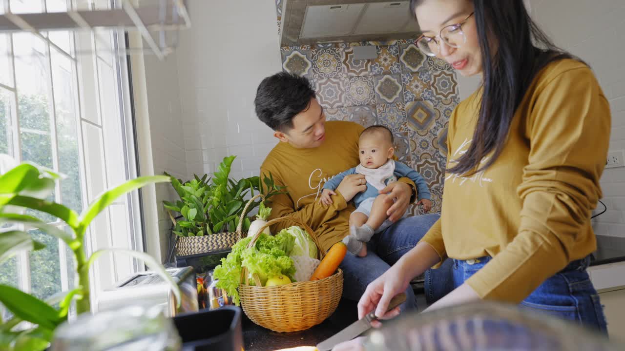 一个新成员的亚洲家庭，5个月大的女婴在厨房里一起准备早餐，父亲抱着女儿看母亲做三明治。视频下载
