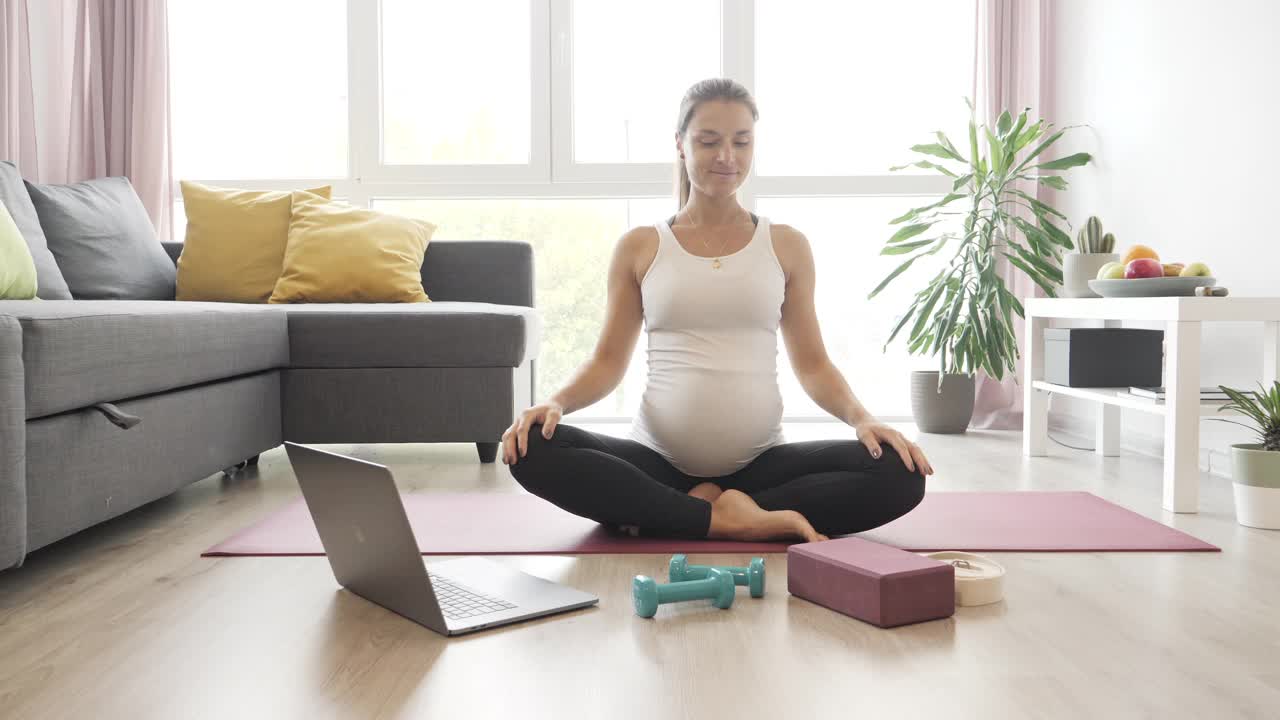 美丽的孕妇在家里用笔记本电脑练习瑜伽。视频素材