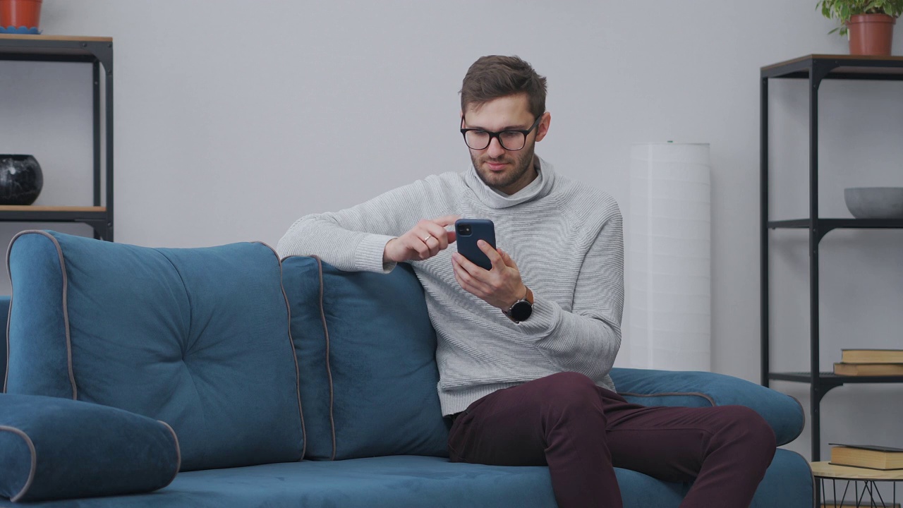 一个戴眼镜的年轻人正在家里用智能手机上网，独自坐在客厅的蓝色沙发上，男性肖像视频素材