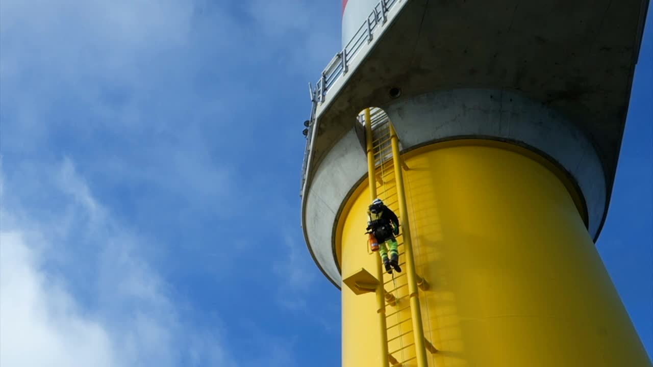 绳索进出技术员(手工高工，工业攀登者)爬上最大的风力涡轮机上的梯子。戏剧性的天空和身后旋转的叶片。视频素材