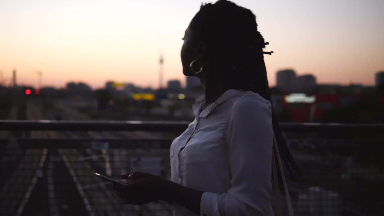年轻女子拿着智能手机看向别处，而行走在城市的人行桥在日落视频素材