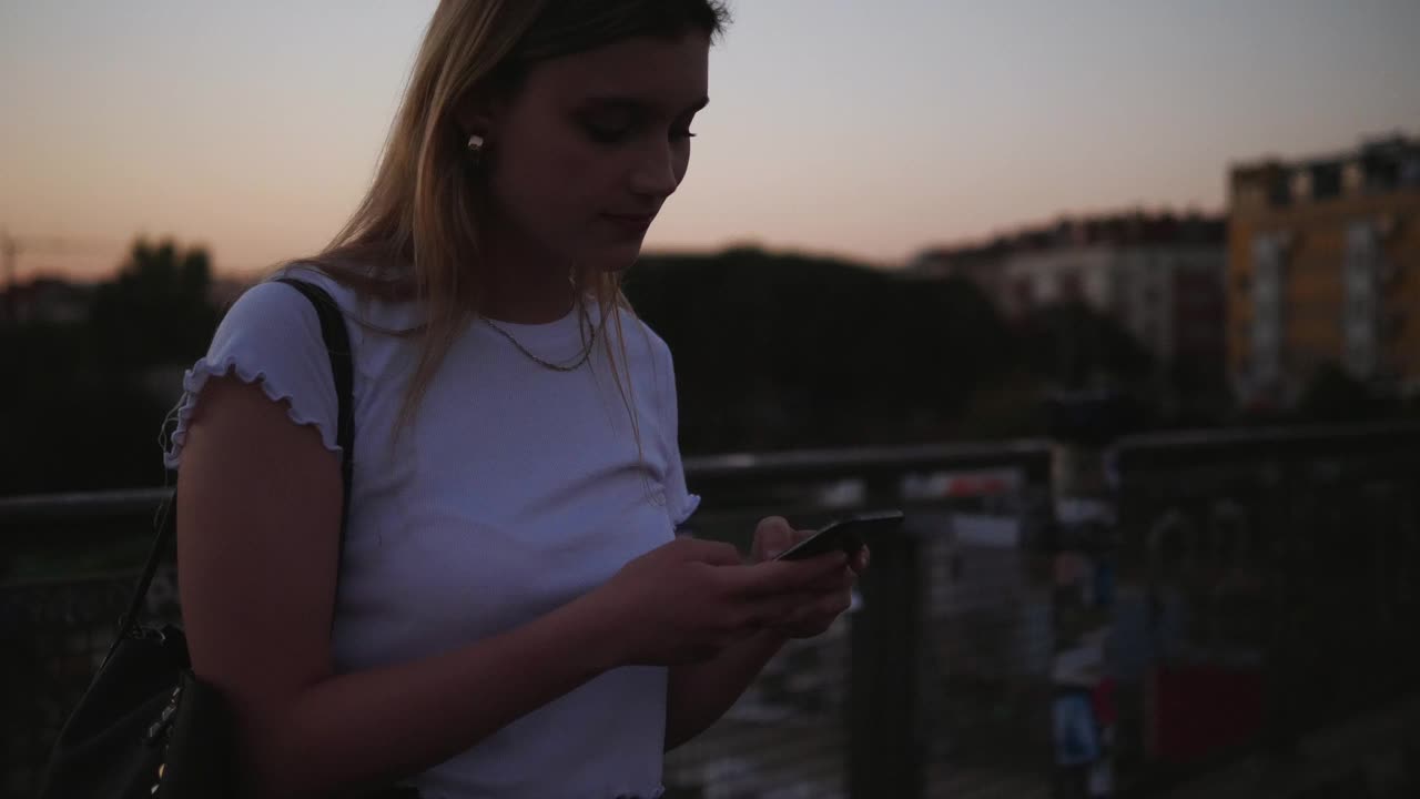 手持拍摄的年轻女子通过智能手机在城市的人行桥上发短信的天空在日落视频素材