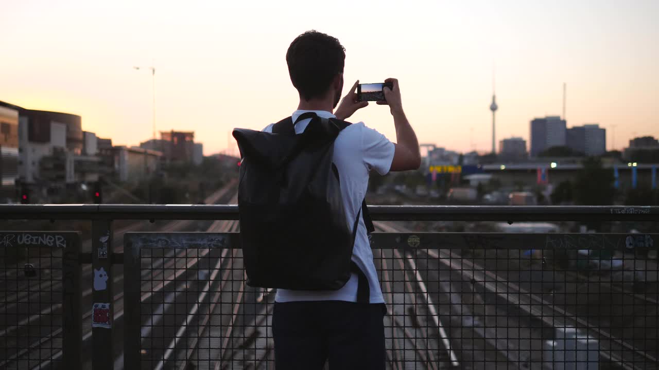 年轻男性游客在日落时站在人行桥上拍摄城市的后视图视频下载