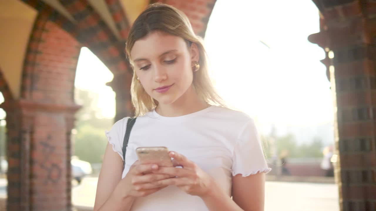 年轻的金发女人微笑着用智能手机发短信，站在城市的人行道上视频素材