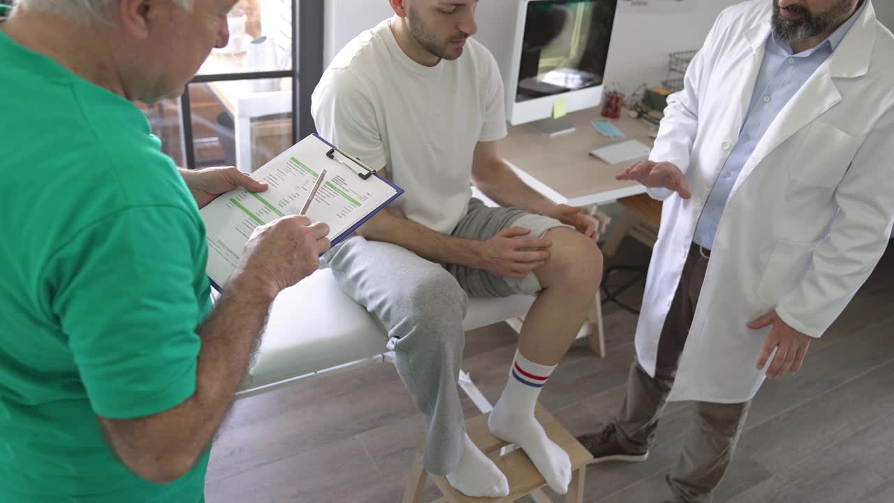 理疗师和他的助手在医生的办公室里与老病人谈论他的膝盖受伤视频素材