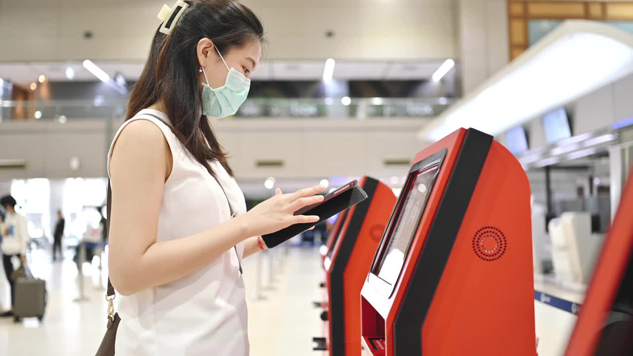 在泰国曼谷，亚洲商务女乘客在机场自检时的广角镜头，手持护照和平板电脑准备登记登机，戴着口罩安全出行视频下载
