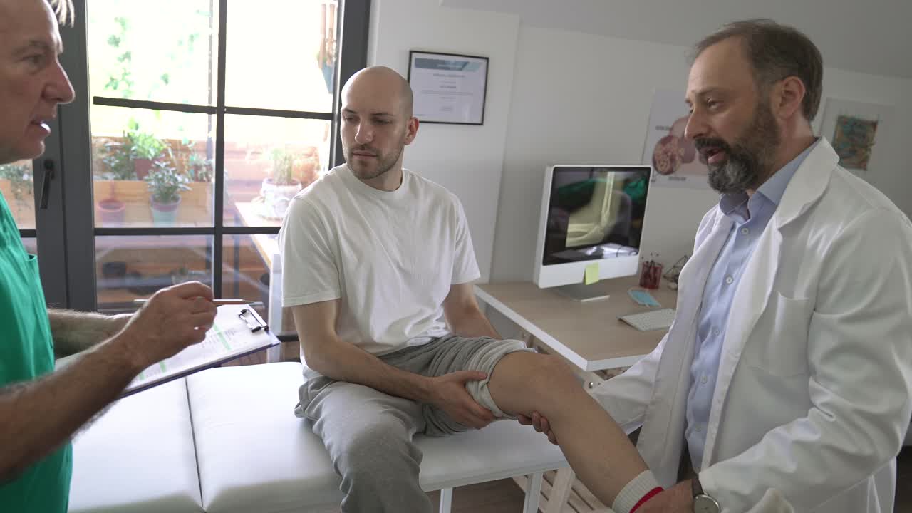 理疗师和他的助手在医生的办公室里与老病人谈论他的膝盖受伤视频下载