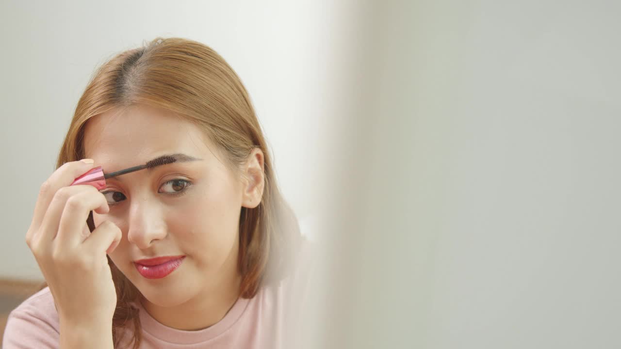 一个年轻的亚洲女人做化妆刷眉毛和看镜子的特写视频下载