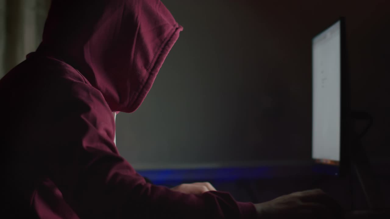 一名戴着兜帽的不明身份男子晚上坐在电脑显示器前，破解密码，发起网络攻击视频素材
