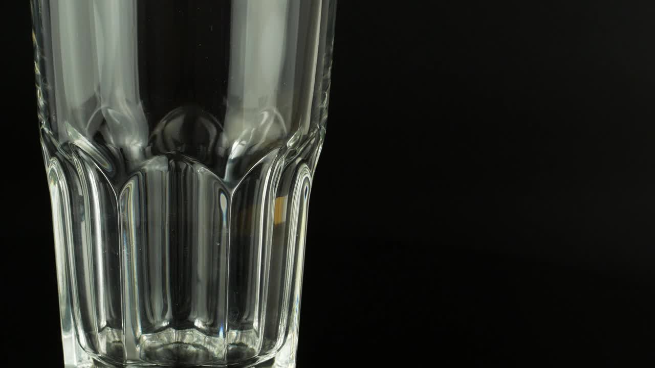 在黑色背景下，冰块落入一个空的透明玻璃杯。4k视频特写玻璃和冰的饮料。视频素材