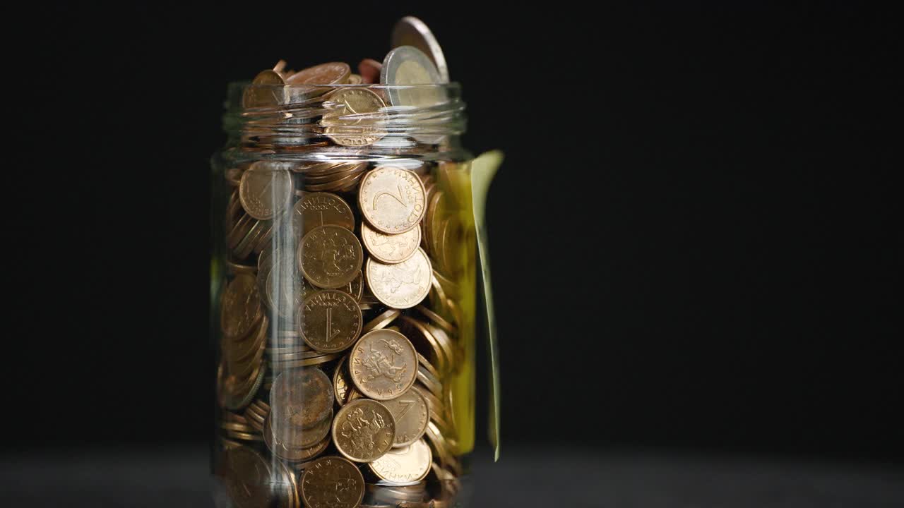 旋转玻璃罐装满硬币和两欧元硬币与粘贴的加密投资文本黑色背景，慢动作视频下载
