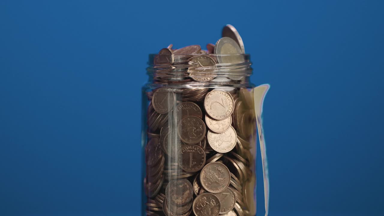装有硬币和两枚欧元硬币的旋转玻璃罐，上面粘贴有加密货币投资文字，蓝色背景，慢动作视频素材