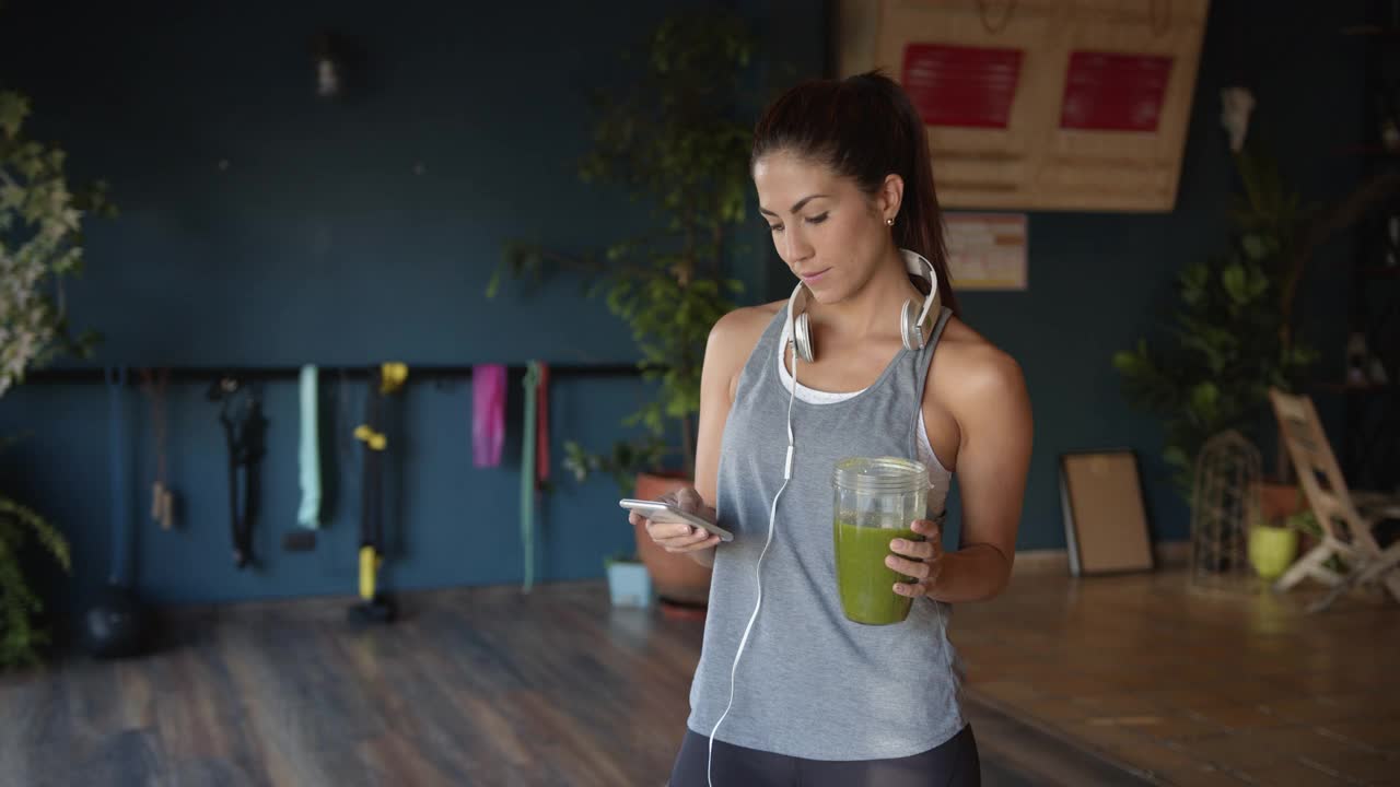 一名年轻的拉丁美洲女子在健身房喝着果汁，脖子上戴着耳机查看智能手机上的信息视频素材