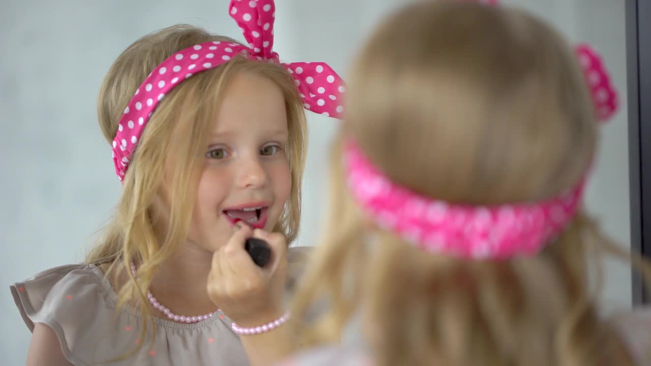 可爱的小女孩涂着红色的口红，化妆，看着镜子微笑。孩子一边玩一边用口红照镜子。小时尚达人化妆，涂她的嘴唇4 K视频素材