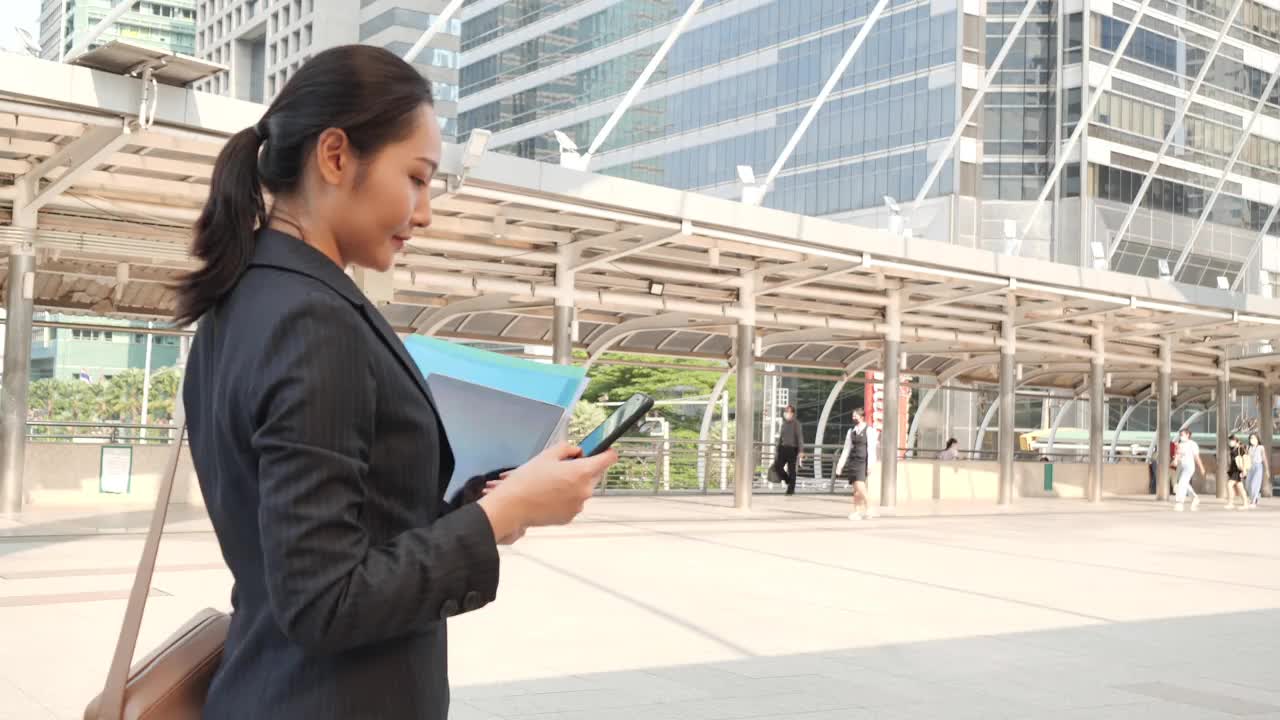 可爱的亚洲女商人一边走一边用手机在城市建筑里。自信和快乐的女人享受工作生活- 4K高质量的镜头视频素材