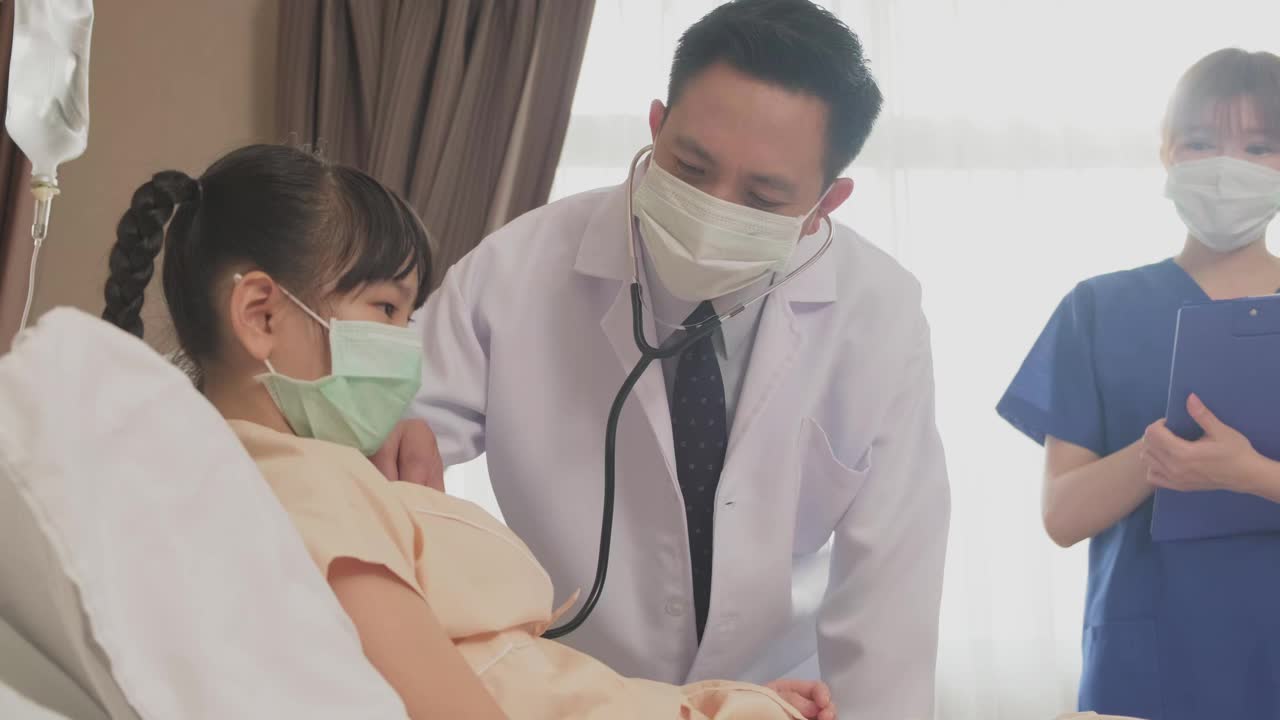 亚洲医生在康复室的床上用听诊器测量小孩的心率。冠状病毒大流行期间，戴口罩的男女检查女童。年轻的护士和孩子说话。视频素材