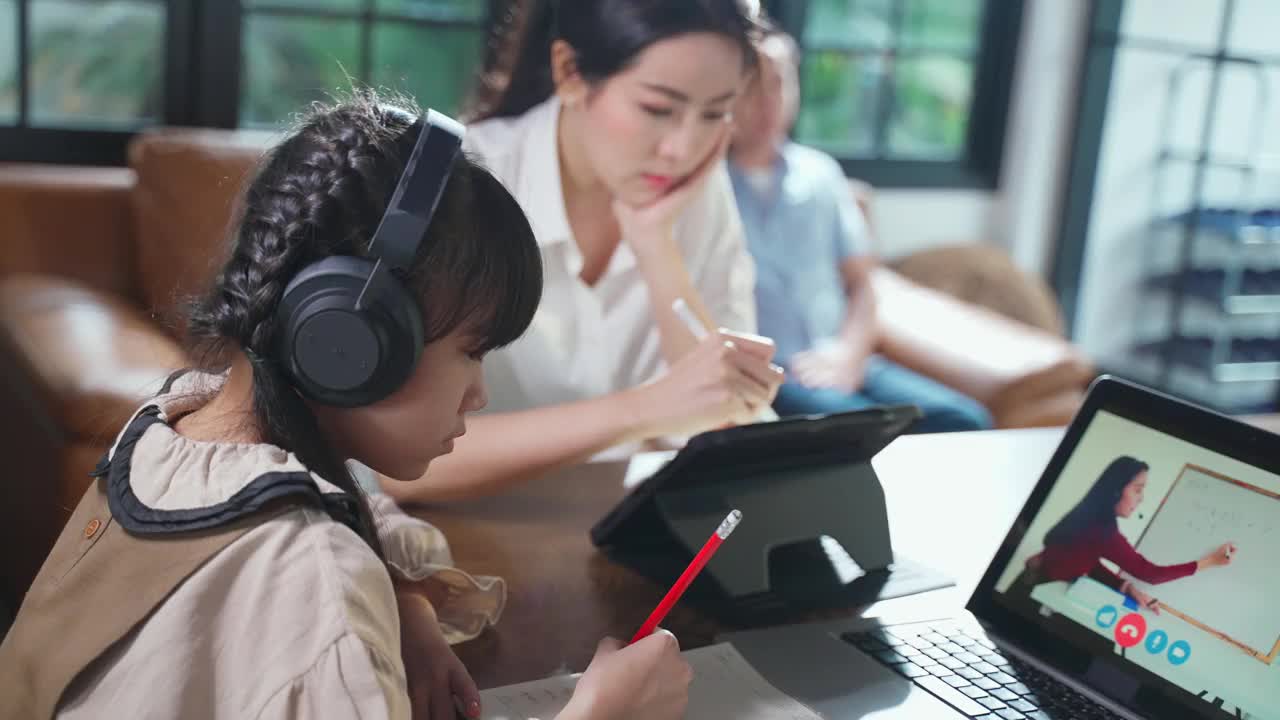 由于冠状病毒大流行，亚洲小女孩通过数字远程互联网会议从学校老师那里学习在线课程。孩子看电脑笔记本电脑屏幕与母亲的协助。视频素材