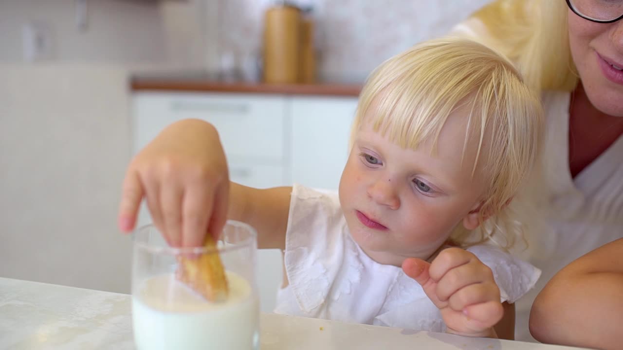 一个金发小女孩的肖像和可爱的笑脸坐在家里厨房，吃饼干与一杯牛奶。孩子在牛奶里蘸饼干，慢动作视频下载
