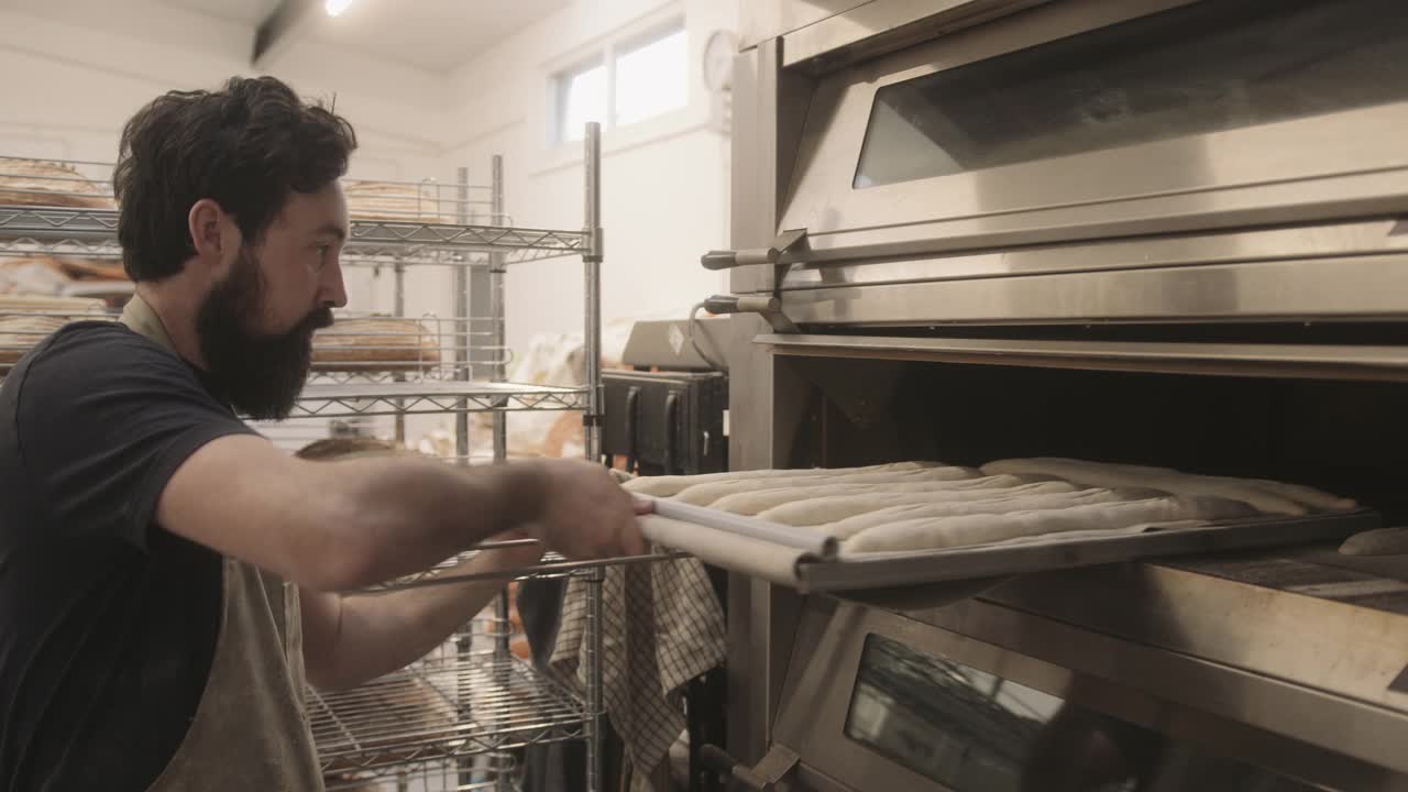 长胡子的男面包师正在把手工法棍放在烤箱里，在工业面包房工作视频素材