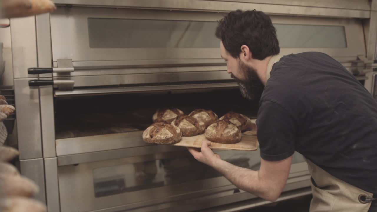 长着胡子的男面包师，穿着围裙从烤箱里取出热烤法棍面包视频素材