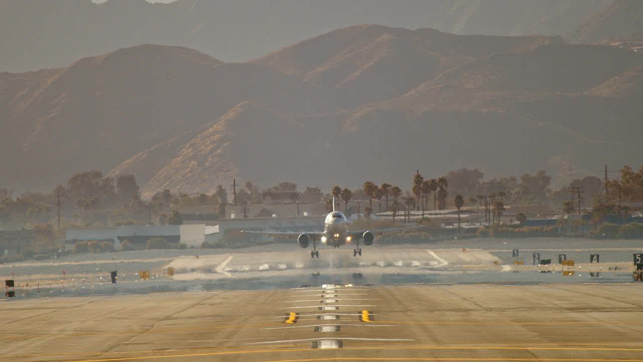 极端长镜头的客机降落在机场跑道上，背景是棕榈树和沙漠山脉，滑行的飞机轮胎喷射白色烟雾降落视频素材