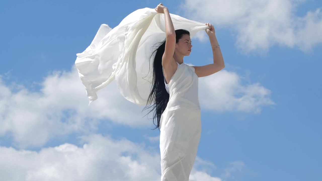 山顶上站着一位美丽的黑发长发女子，她穿着飘逸的白色衣服，映衬着天空。天使存在的幻影。200帧/秒的慢动作视频下载