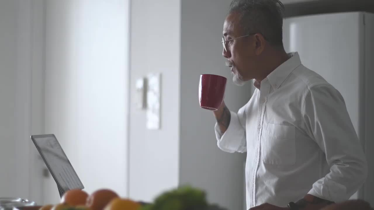 胡子花白头发的亚洲华人老人在厨房柜台边喝着咖啡边用着平板电脑视频素材