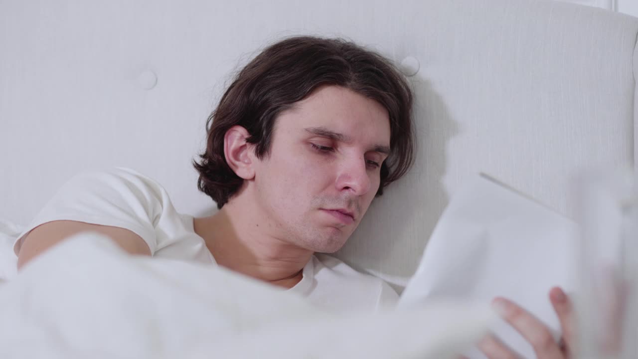特写的疲惫的年轻人睡着了，在晚上阅读。英俊的专注的白种人拿着书睡在家里的床上。爱好和生活方式视频素材