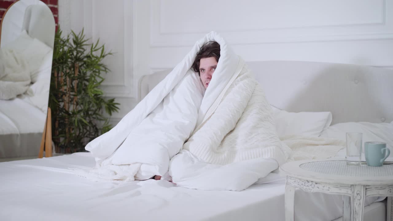 生病的年轻人发烧坐在床上裹着白色的羽绒被。一个生病的高加索人在家里的卧室里高温。健康舒适视频素材