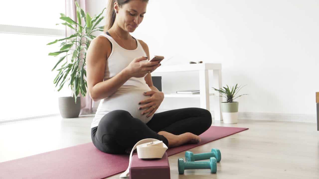 美丽的孕妇一边练瑜伽一边玩手机。视频素材