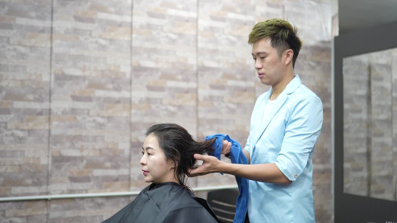 亚洲华人理发师，在他的沙龙里给一个女人吹头发。视频素材